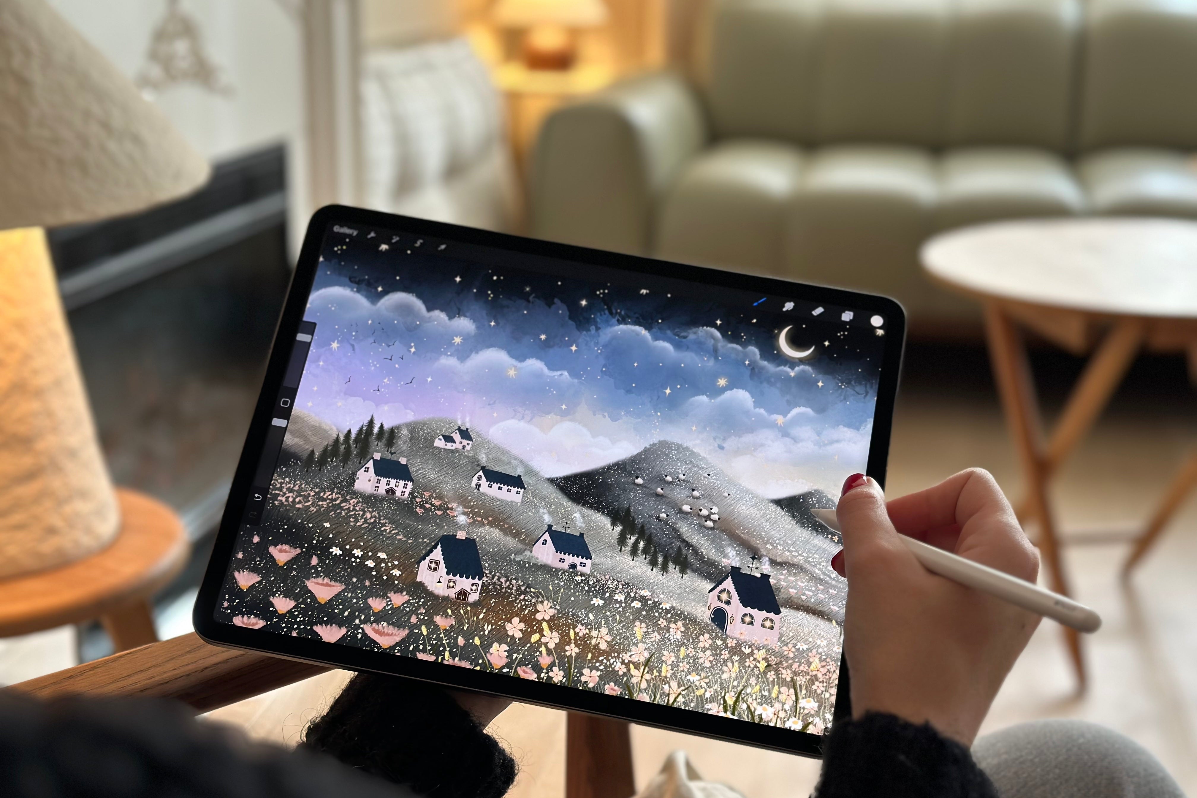 Person holding a tablet with a drawing of houses on a mountain landscape in a cozy living room.